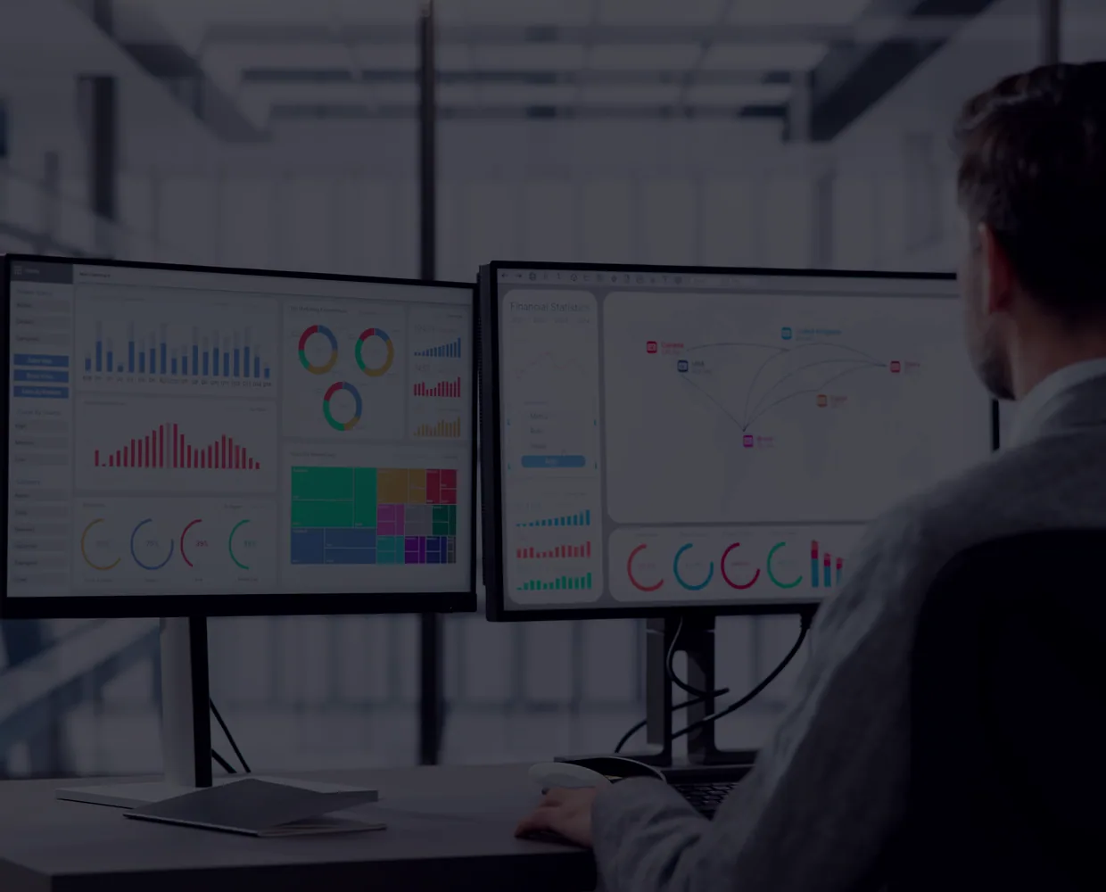An analyst at two monitors of dashboards in an open office.