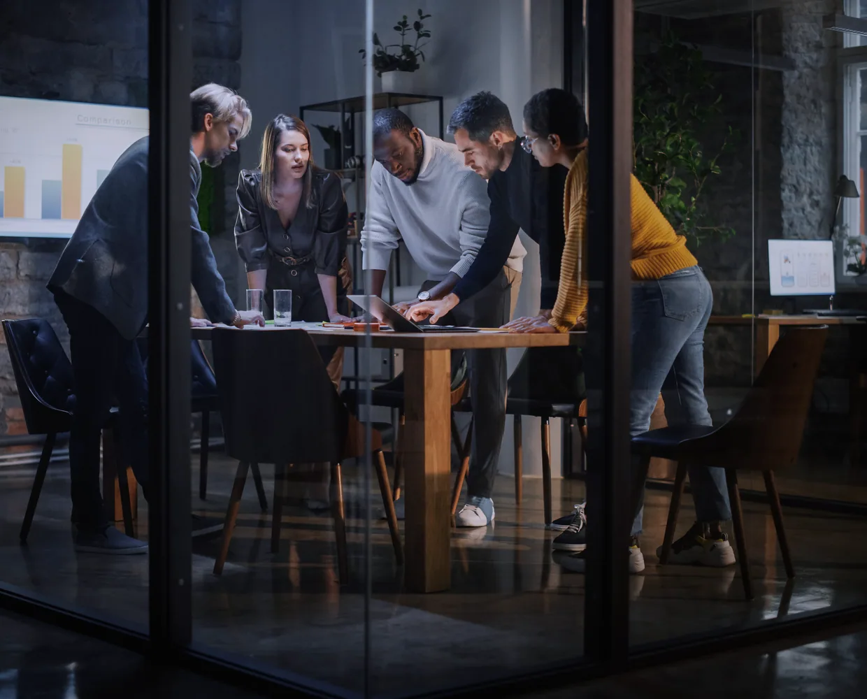 A leadership team around a table in a glass-walled meeting room at night, working through a plan together.